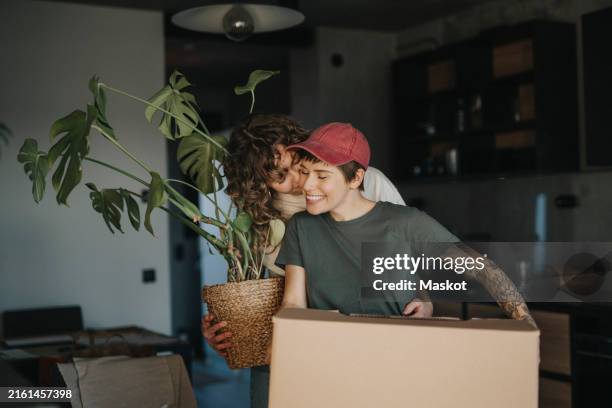 smiling lesbian woman holding houseplant kissing girlfriend carrying cardboard box during relocation - mudar de casa imagens e fotografias de stock