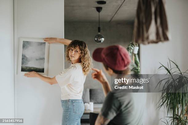 lesbian woman taking advice from girlfriend while hanging frame on wall at home - decorar fotografías e imágenes de stock