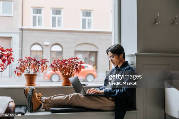 businessman working on laptop while sitting on window sill at office - window sill stock pictures, royalty-free photos & images