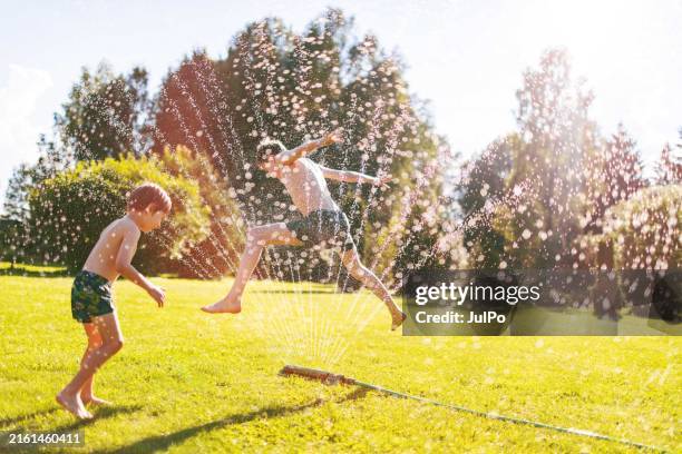 two boys jumping over sprinklers - sprinkler stock pictures, royalty-free photos & images