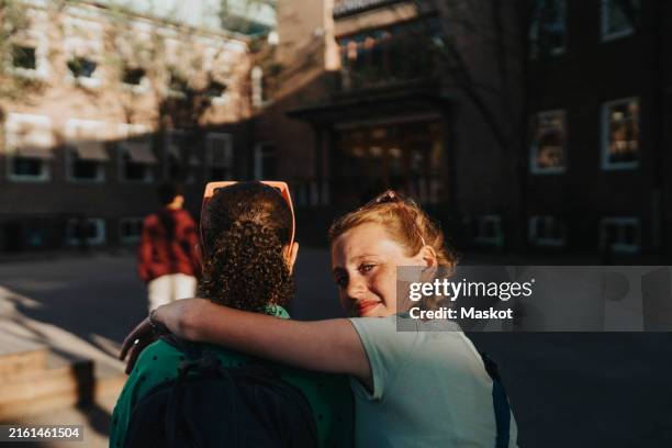 portrait of smiling girl with arm around friend looking over shoulder while walking at school campus - arm umlegen stock-fotos und bilder