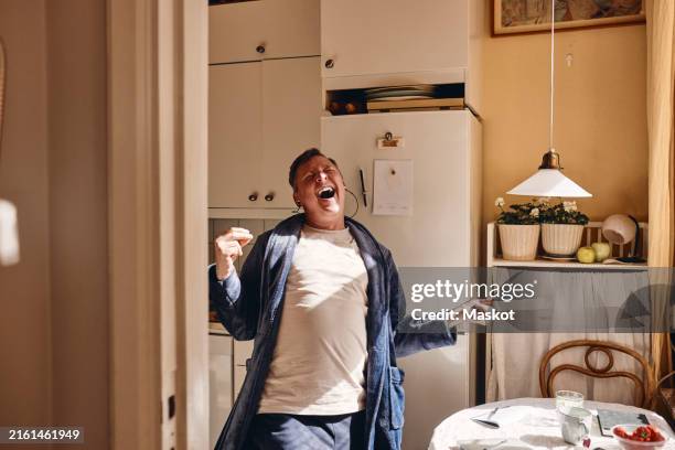 cheerful mature man listening to music while standing in kitchen at home - schreeuwen stockfoto's en -beelden