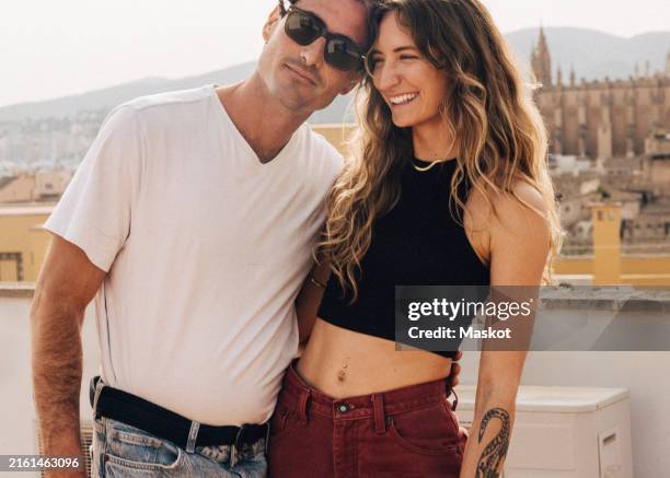 smiling woman standing with boyfriend wearing sunglasses on building rooftop - boyfriend stock pictures, royalty-free photos & images