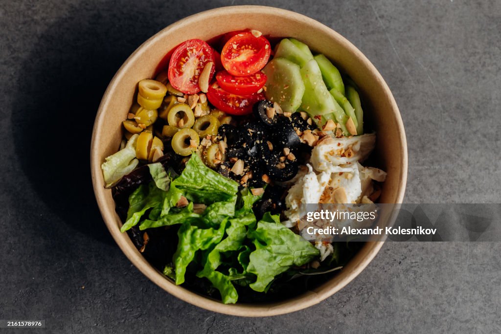 Delicious food bowl with Stracciatella cheese, olives, lettuce, vegetables and sauce in disposable eco-friendly packaging. High angle view, flat lay