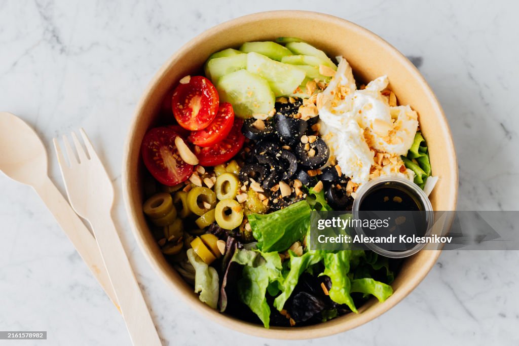 Healthy food bowl with Stracciatella cheese, olives, lettuce, vegetables and sauce in disposable eco-friendly packaging. Take out balanced food. High angle view, flat lay