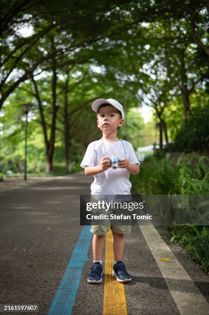 young boy holding camera on a pathway under lush green trees, neutral mood. - body camera stock pictures, royalty-free photos & images