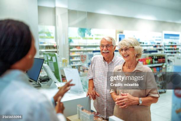 cheerful senior couple consulting about medicine with pharmacy worker at counter - apotheek stockfoto's en -beelden