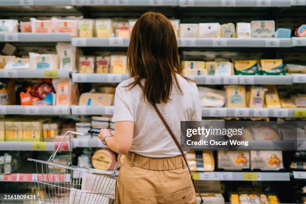 rear view of young woman carrying shopping basket, choosing cheese, standing in front of produce aisle in supermarket - stormarknad bildbanksfoton och bilder