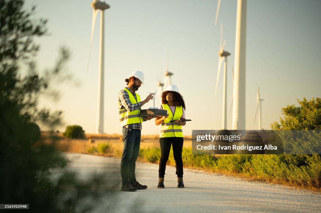 Two engineers discussing about a wind turbine model on a wind farm at sunset
