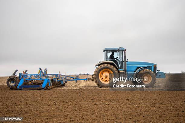 blue tractor plowing a field on a cloudy day - tractor stockfoto's en -beelden