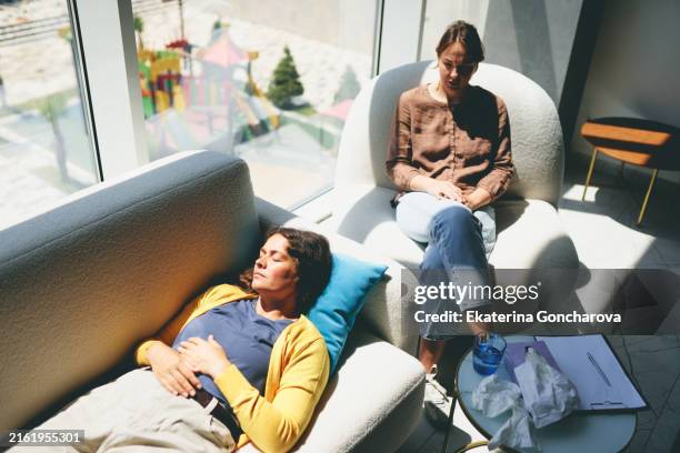 woman lying on a sofa with eyes closed during therapy. therapist taking notes with clipboard. bright room with large windows. - hypnosis stock pictures, royalty-free photos & images