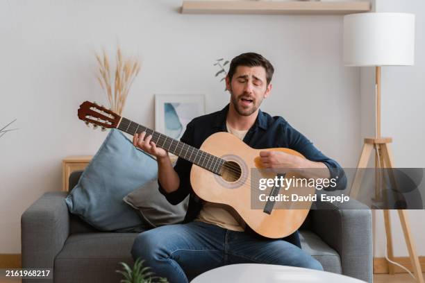 musician playing acoustic guitar and singing at home on sofa - akustikgitarre stock-fotos und bilder