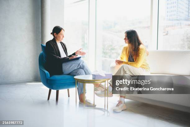 two women having a serious conversation in a bright room with large windows. one woman takes notes while the other listens. - therapie stockfoto's en -beelden