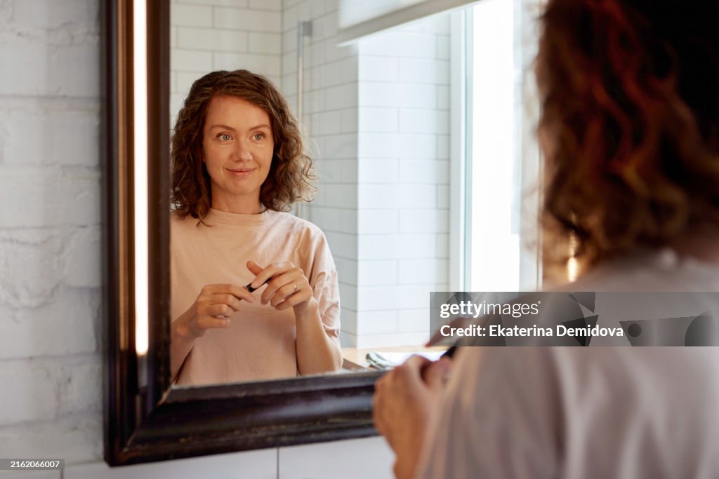 In a cheerful scene, a woman happily applies makeup in a welllit bathroom, admiring her reflection in the mirror