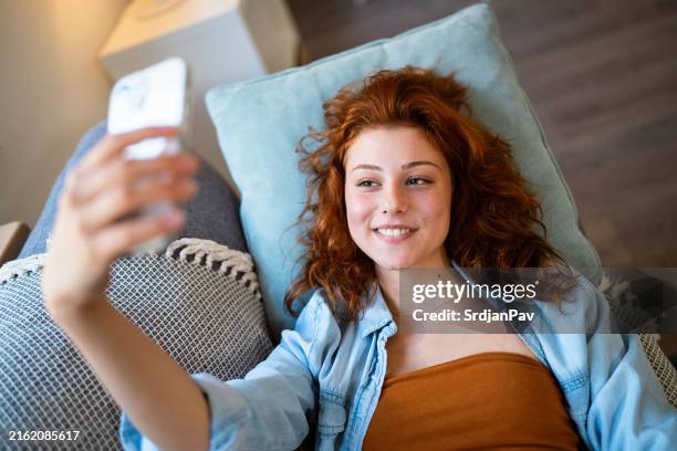 redhead caucasian woman lying on the sofa and taking selfie with mobile phone - selfie girl stockfoto's en -beelden