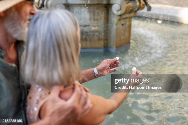 senior couple throwing coins into fountain, making a wish. pensioners enjoying hot summer in the city. staycation concept. - coin fountain stock pictures, royalty-free photos & images