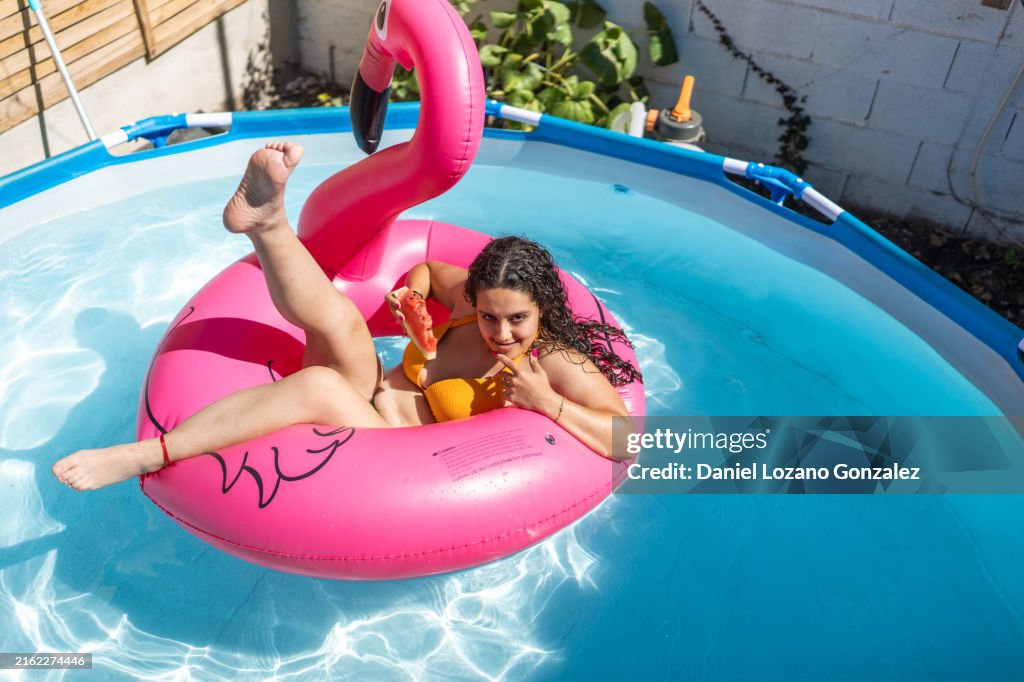Young woman relaxing on inflatable flamingo in swimming pool eating watermelon