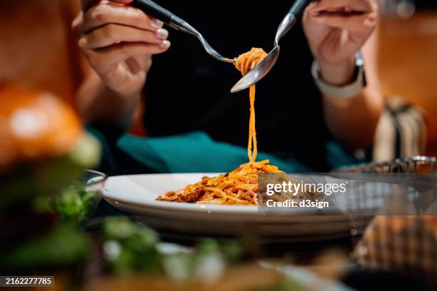 close-up, mid-section of young asian woman sitting at dining table holding cutleries, twirling freshly served spaghetti bolognese and enjoying her meal in restaurant. italian food. dining out. people, food and lifestyle - pasta fotografías e imágenes de stock