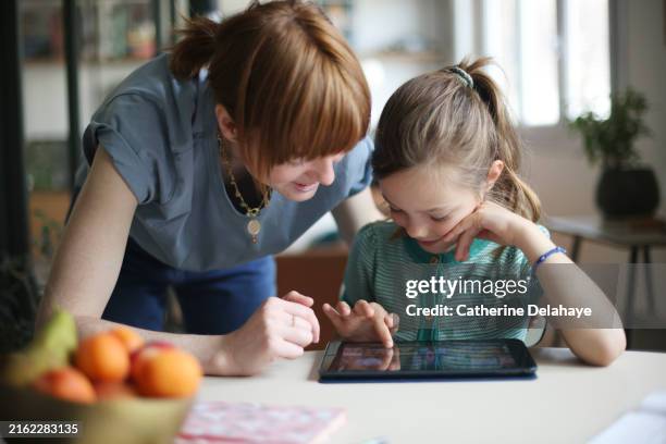 a mother and her daughter looking at a tablet on the kitchen table at home - homemaker stock pictures, royalty-free photos & images