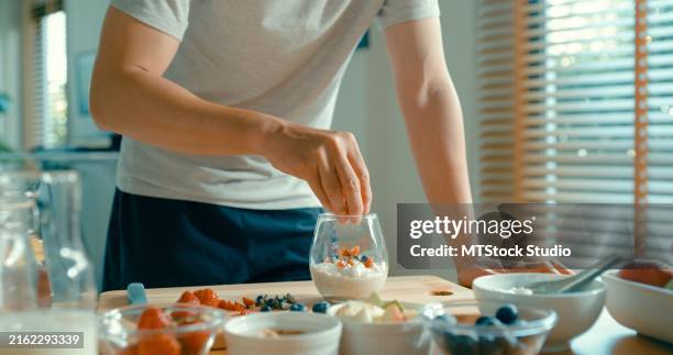 nahaufnahme eines jungen asiatischen mannes, der joghurt- und müslifrühstück in einem modernen haus zubereitet. diät und gesunde ernährung morgenroutine. - superfood stock-fotos und bilder