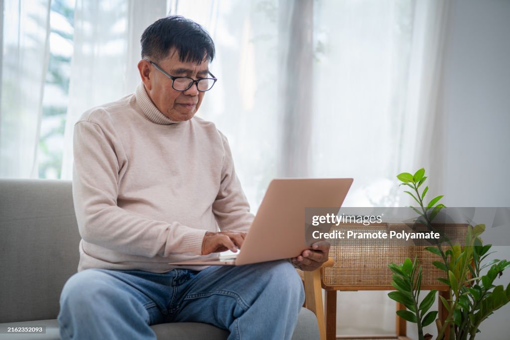 Asian happy senior man using laptop on sofa at home.