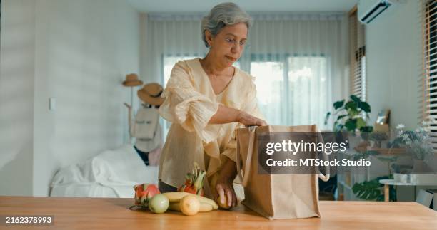 mujer asiática madura desempacando bolsas de papel de comestibles frescos en casa. vida sostenible. - bolsa reutilizable fotografías e imágenes de stock