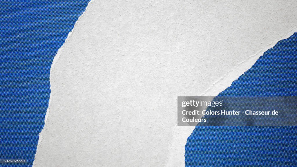 Close-up of a torn blue poster, printed in offset (ink dots visibles) and pasted on a white wall in Paris, France. Rough texture. Natural light and colors.