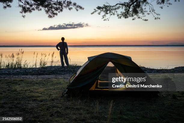 tourist enjoys sunset in camp with tents on lake coast. slow life and meditating for nature lover. mindfulness concept - wilderness survival stock pictures, royalty-free photos & images