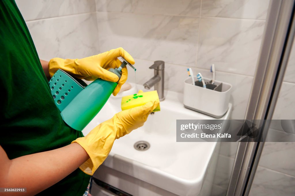 A hand in protective rubber gloves is seen wiping and cleaning the sink in the bathroom