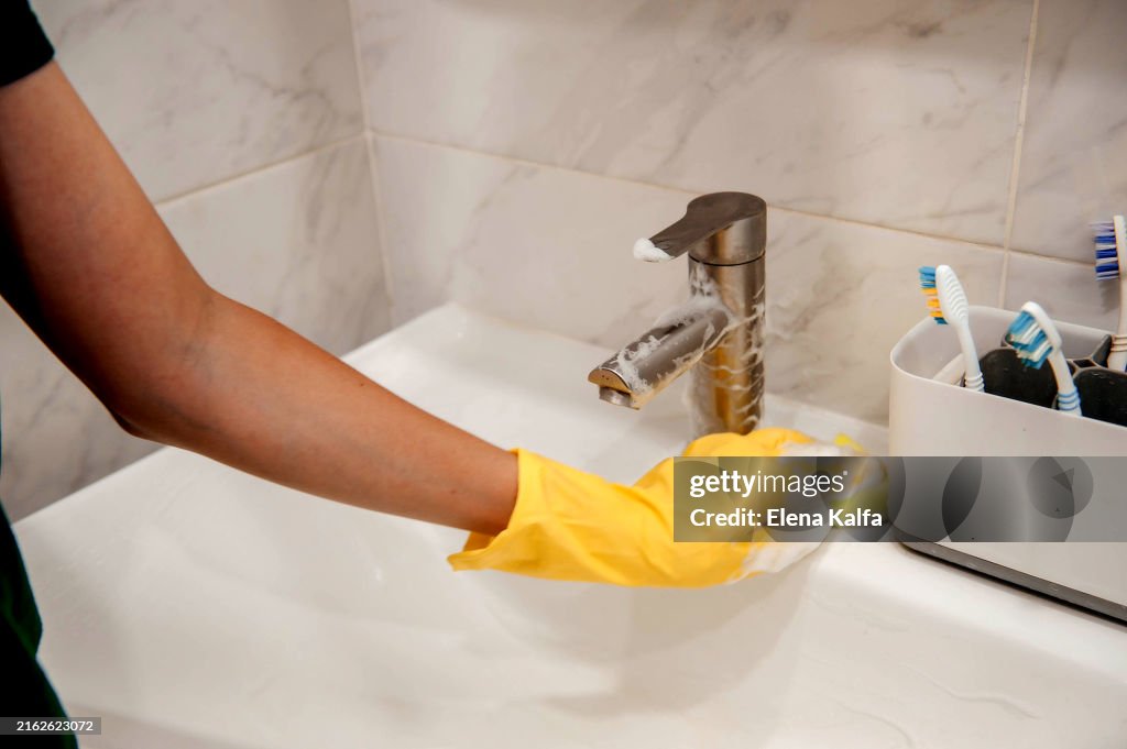 A hand in protective rubber gloves is seen wiping and cleaning the sink in the bathroom