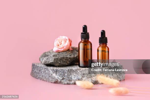 two cosmetic dropper bottles on stones with roses and dried flowers on a soft, solid pink background with copy space. conceptual still life, modern composition. front view, studio shot. - jojoba stock pictures, royalty-free photos & images