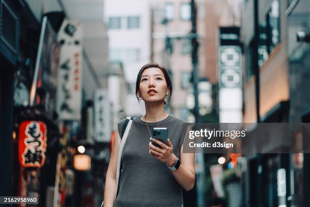 low angle shot of young asian female traveller using smartphone to navigate city streets in japan. exploring around the famous travel spots. wanderlust. travel, vacation, exploration and discovery concept. lifestyle and technology - local landmark stock pictures, royalty-free photos & images