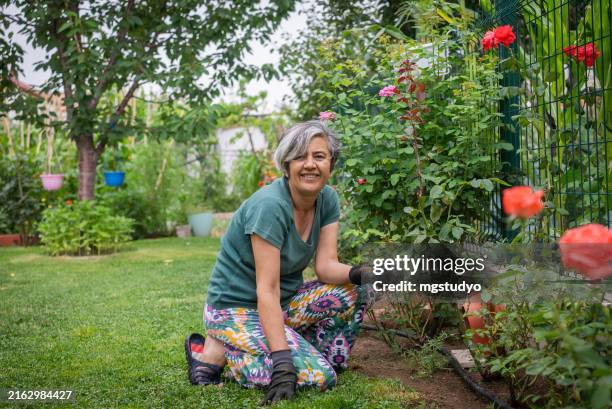 mature woman smiling at the camera while tending to flowers in the garden - gardening glove stock pictures, royalty-free photos & images