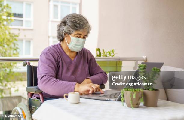 disabled senior woman wearing a face mask sitting in a wheelchair using laptop on the balcony - weakness stock pictures, royalty-free photos & images