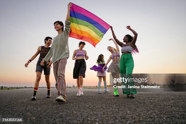 view of a group of young people holding a rainbow flag together, half-dancing and smiling along a path - pride month stock pictures, royalty-free photos & images