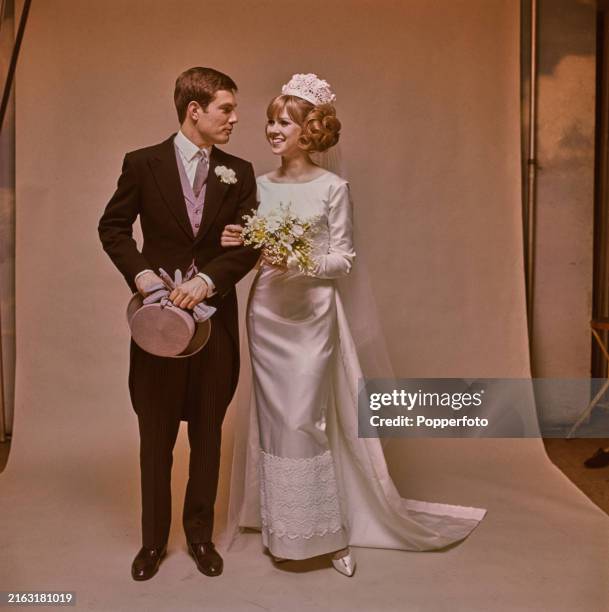Posed studio portrait of a bride and groom dressed for their wedding day, the bride wears a full length white satin gown with train, headdress and...