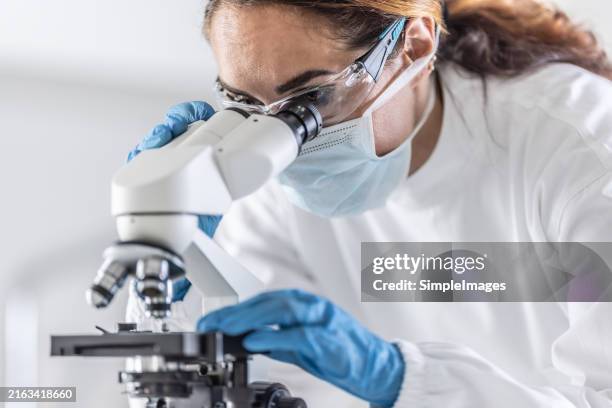 serious handsome handsome scientist sitting at microscope looking and examining sample of virus or vaccine - mikroskop stock-fotos und bilder