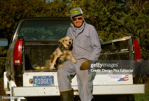 Agriculture - A farmer in coveralls sits on the tailgate of his pickup truck with his dog/Iowa, USA.