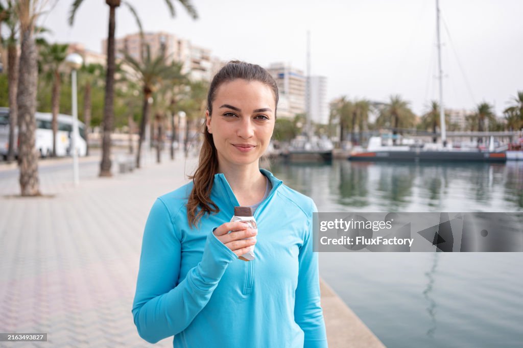 Portrait of a young sportswoman eating a protein bar after running, by the sea