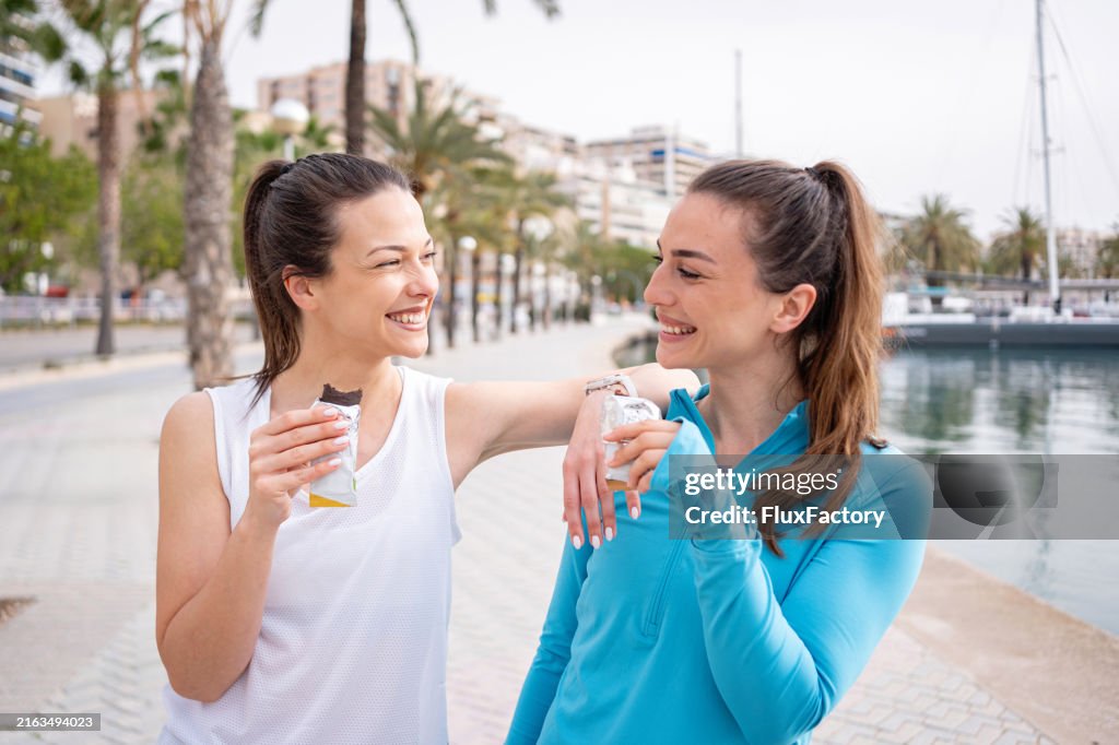 Two female friends eating a protein bar after training on the quayside