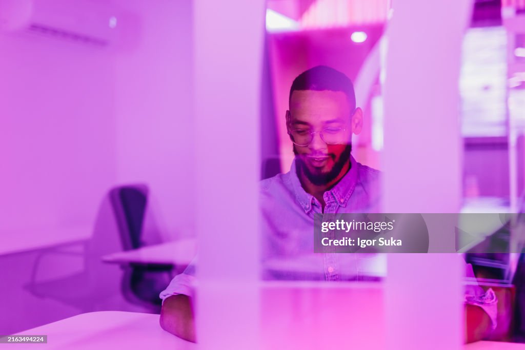 A young man in a shirt and glasses works at a laptop. a man in glasses works in the office in the evening