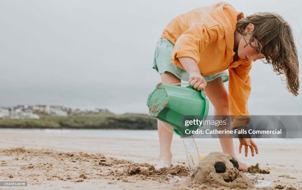 A child pours sea water from her green bucket onto a face she has created in the sand