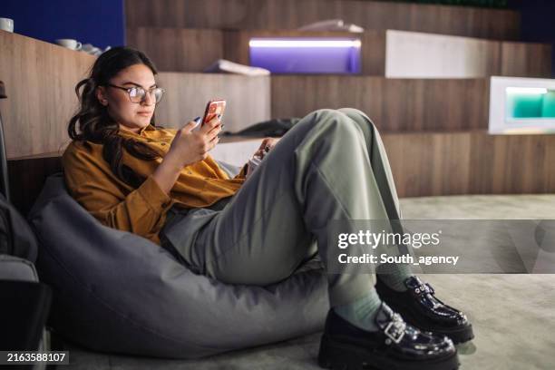 young woman lounging on a bean bag using smartphone in an office. - bean bag stock pictures, royalty-free photos & images