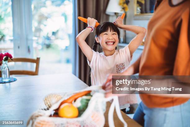 playful young daughter with carrots unpacking groceries with her mother in kitchen at home - cooking vegetables stock pictures, royalty-free photos & images