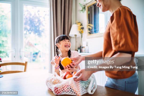 young cheerful girl and her mother unpacking fresh fruits and vegetables together in kitchen - duurzaam consumeren stockfoto's en -beelden