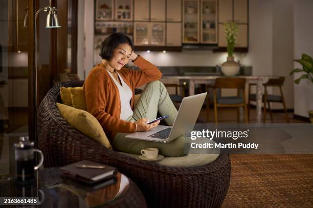 mature asian woman working from home sitting in open plan house, using mobile phone and laptop computer at night - comfortabel stockfoto's en -beelden