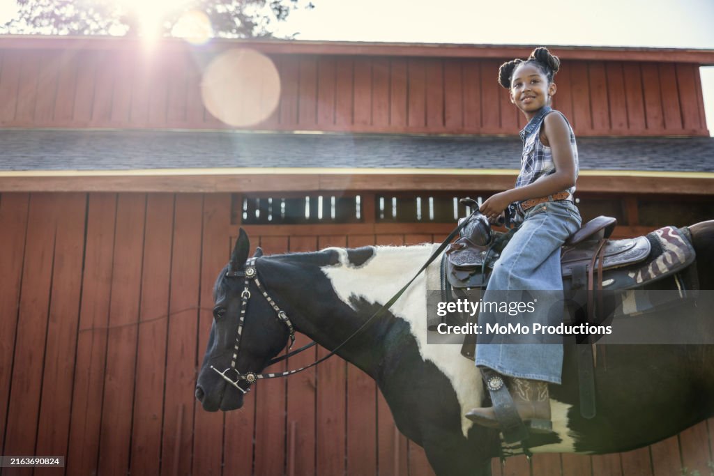 Young girl riding on horse in front of barn