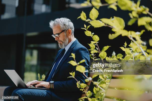 senior businessman working on laptop outdoors in urban garden - lush foliage stock pictures, royalty-free photos & images