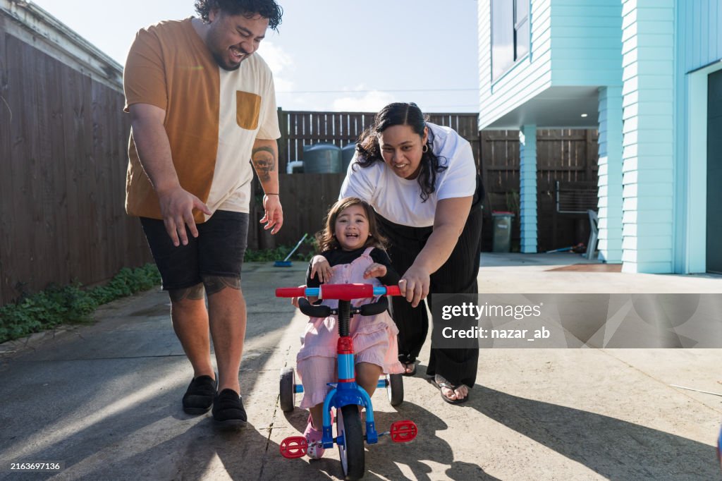 Mom and dad assisting daughter to ride tricycle.