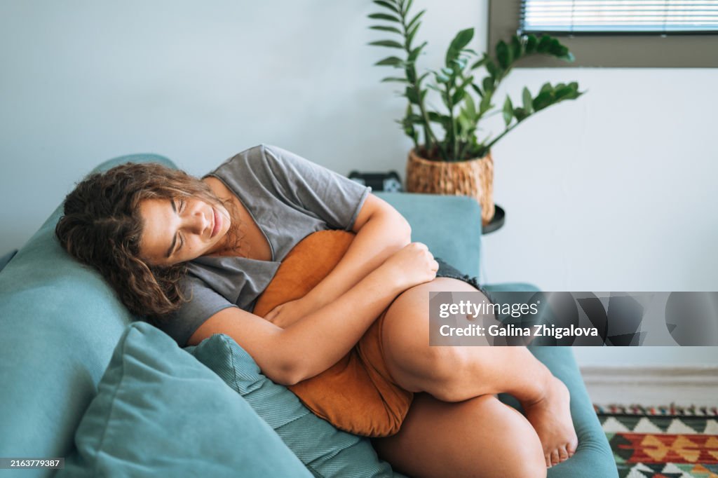 Depressed brunette young woman with pillow in hands sitting on sofa at home